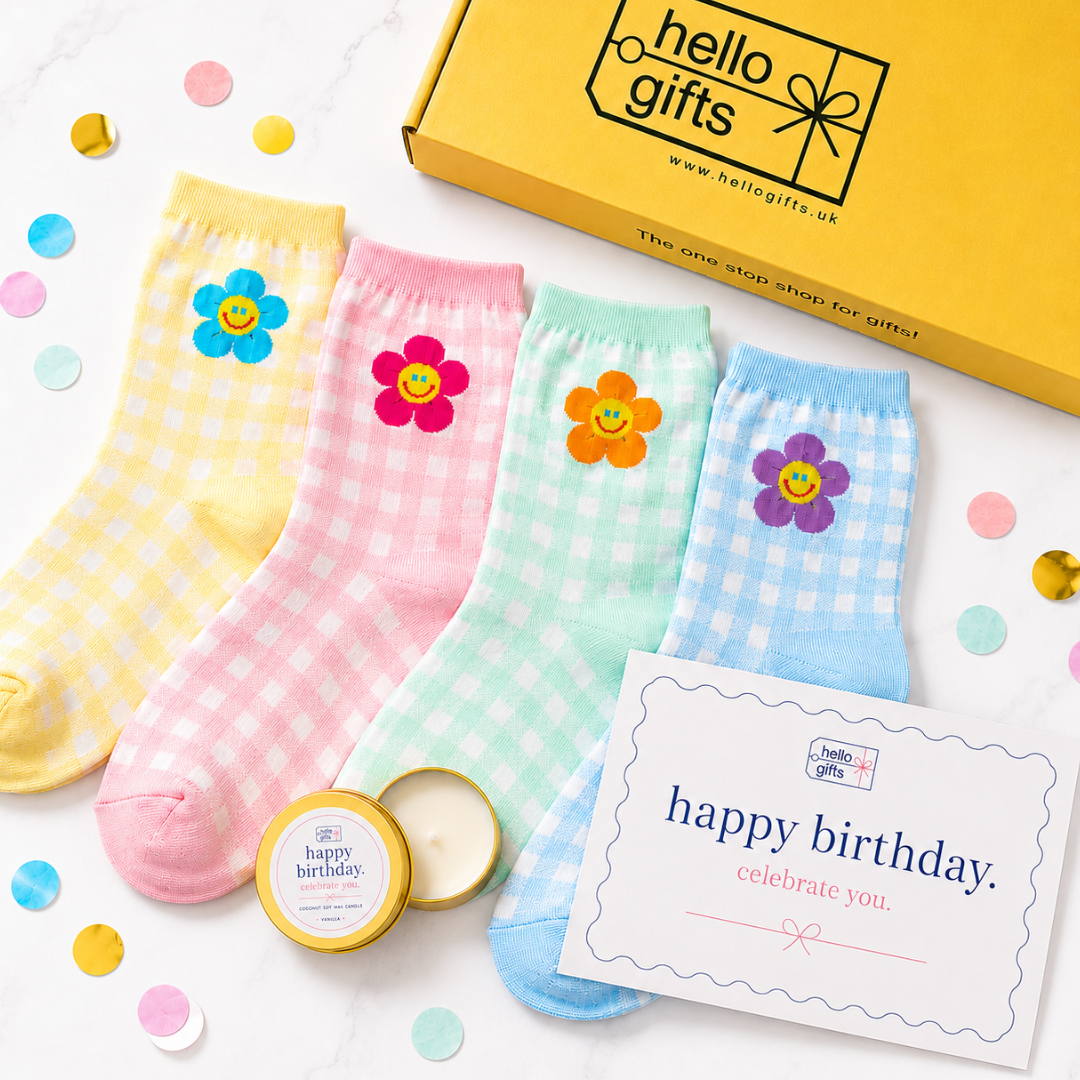Four pastel gingham socks with colorful smiley-face flowers are arranged on a white marble surface with confetti. A yellow Hello Gifts box, a small candle tin, and a birthday card are also visible.