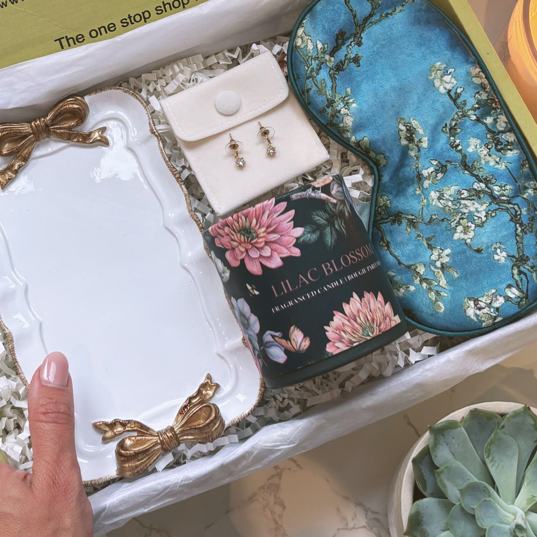 A gift box opened to reveal a white trinket plate with gold bow details, a satin feel eye mask, a pair of sparkle drop earrings, and a blossom-scented candle.