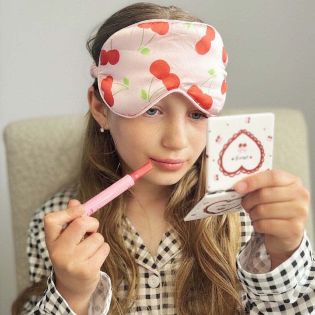 Girl wearing a cherry-patterned sleep mask, holding a mirror and lip balm