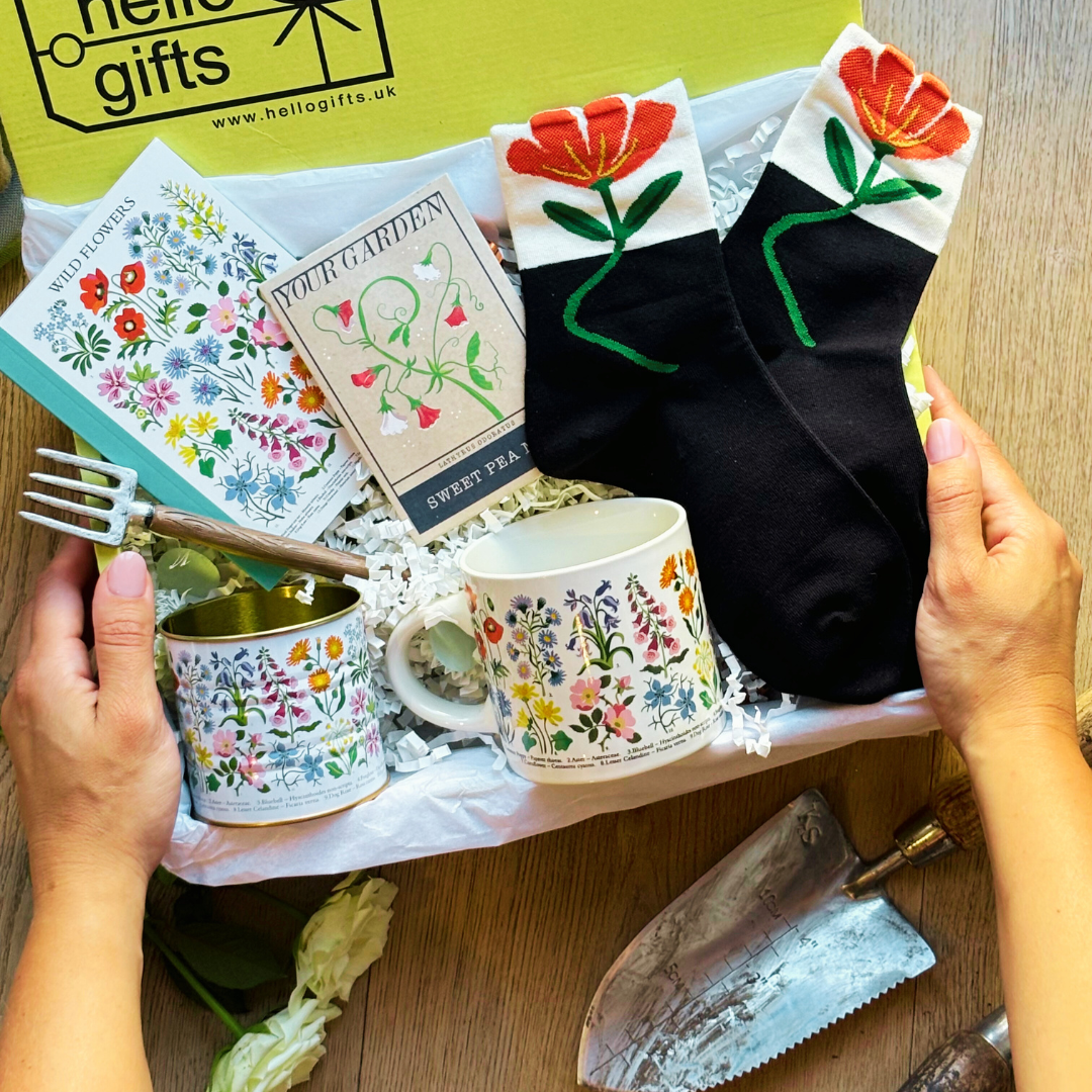 A gift box containing various gardening themed items including a ceramic mug with a floral design, a tin with a floral print, a notebook, a pair of socks with a floral pattern, and a packet of flower seeds, all branded with &