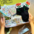 A gift box containing various gardening themed items including a ceramic mug with a floral design, a tin with a floral print, a notebook, a pair of socks with a floral pattern, and a packet of flower seeds, all branded with &
