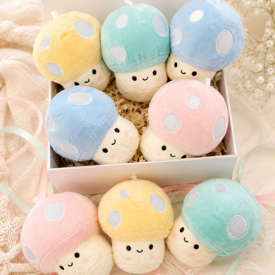 A set of plush mushroom toys in pastel colors, including yellow, mint green, light blue, pink, and white, arranged inside and around a white gift box on a soft beige fabric background.
