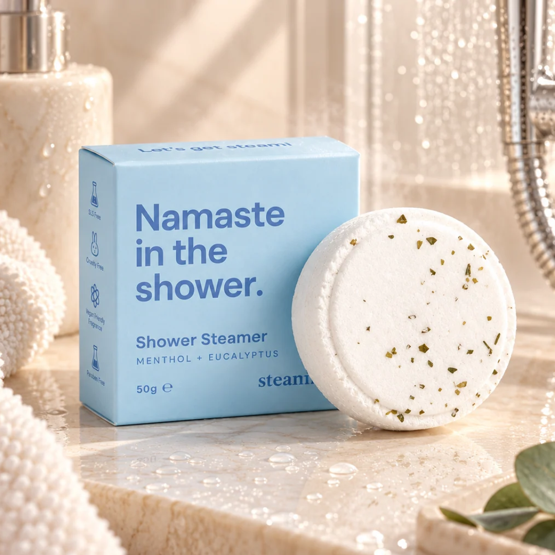 A light blue box of Namaste in the Shower steamers next to a round white bath steamer disc with dried herb specks, set on a marble bathroom counter with a shower head and soap dispenser in the background.