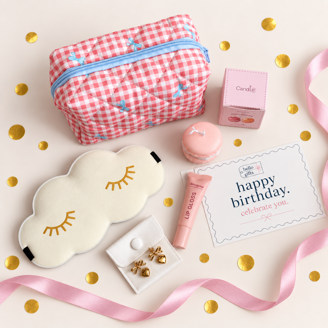 A styled flat-lay of a birthday gift set featuring a pink and white gingham quilted cosmetic pouch with a blue zipper, a cream cloud-shaped sleep mask with gold embroidered eyelashes, a pink macaron, a tube of lip gloss, a small pink box, gold heart earrings on a white card, and a &