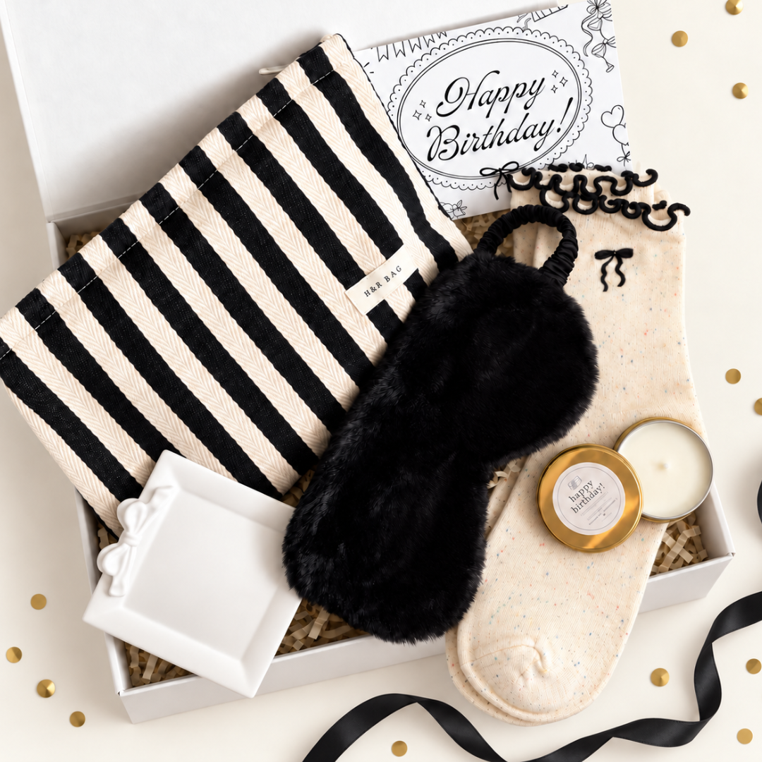 A curated gift box set featuring a black and cream striped bath towel, a black faux-fur sleep mask, cream speckled socks with black embroidery, a white candle in a gold tin, a small white gift box, and a &