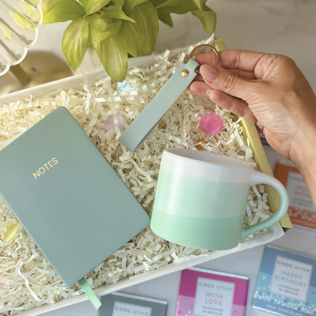 Notebook, mug and keyring gift for her. Hand holding a light green mug with a matching notebook labeled &
