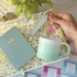Notebook, mug and keyring gift for her. Hand holding a light green mug with a matching notebook labeled &