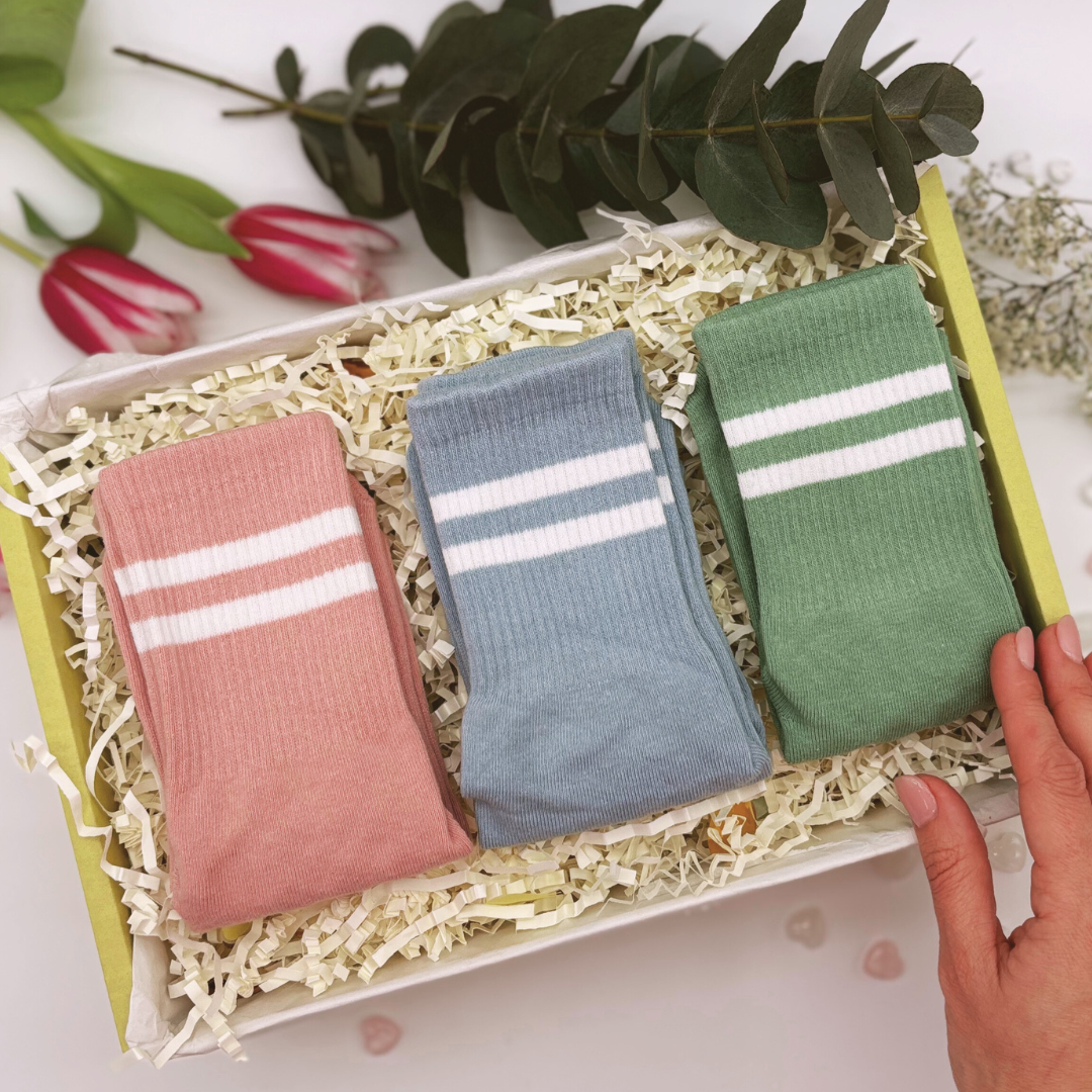 Pilates and yoga socks gift set. A gift box set containing three pairs of pastel-coloured grip socks with white stripes, displayed on a bed of shredded paper. The box is adorned with decorative elements, including flowers and foliage outside.