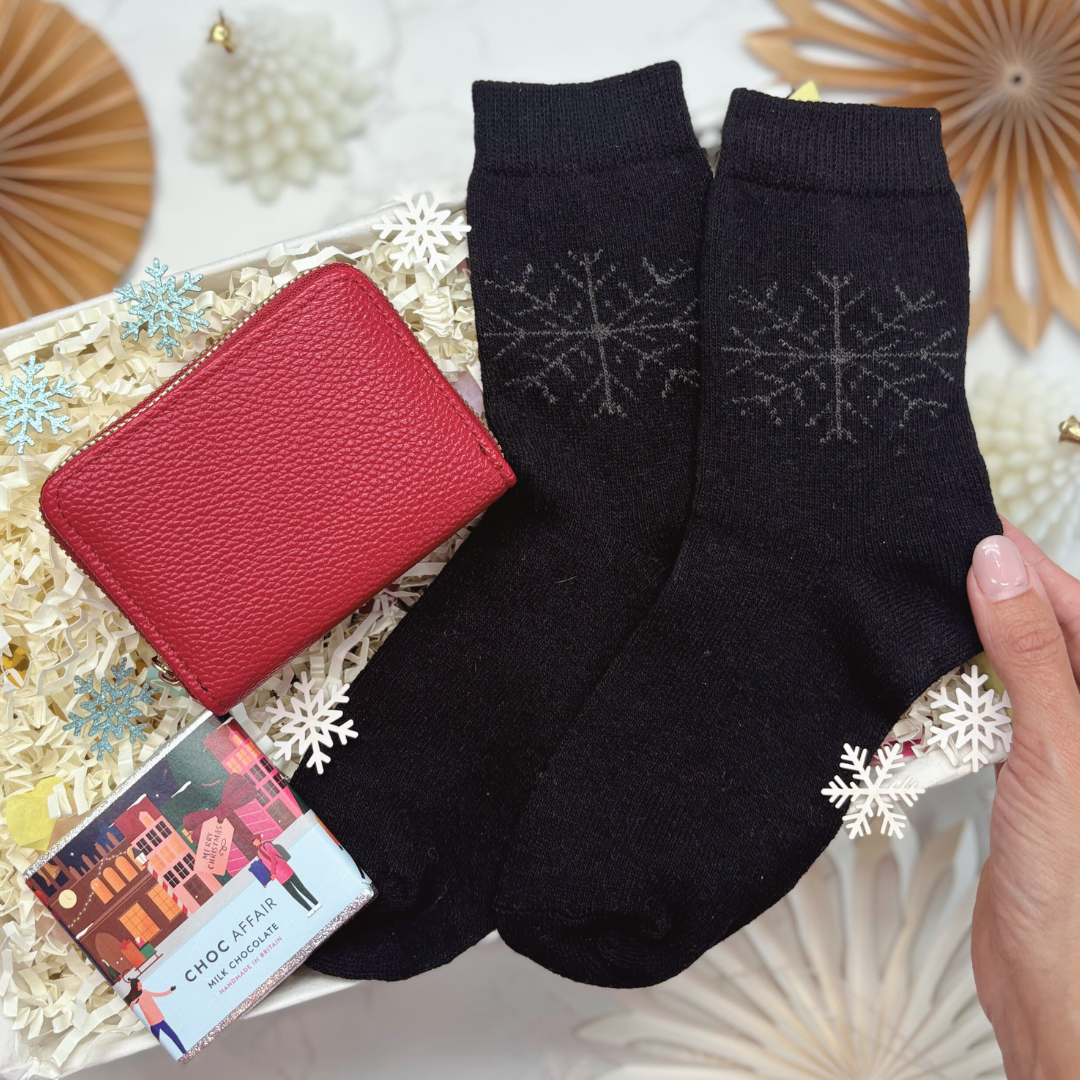 Black socks with snowflake patterns held by a hand, next to a red wallet and chocolate on a decorative gift box.
