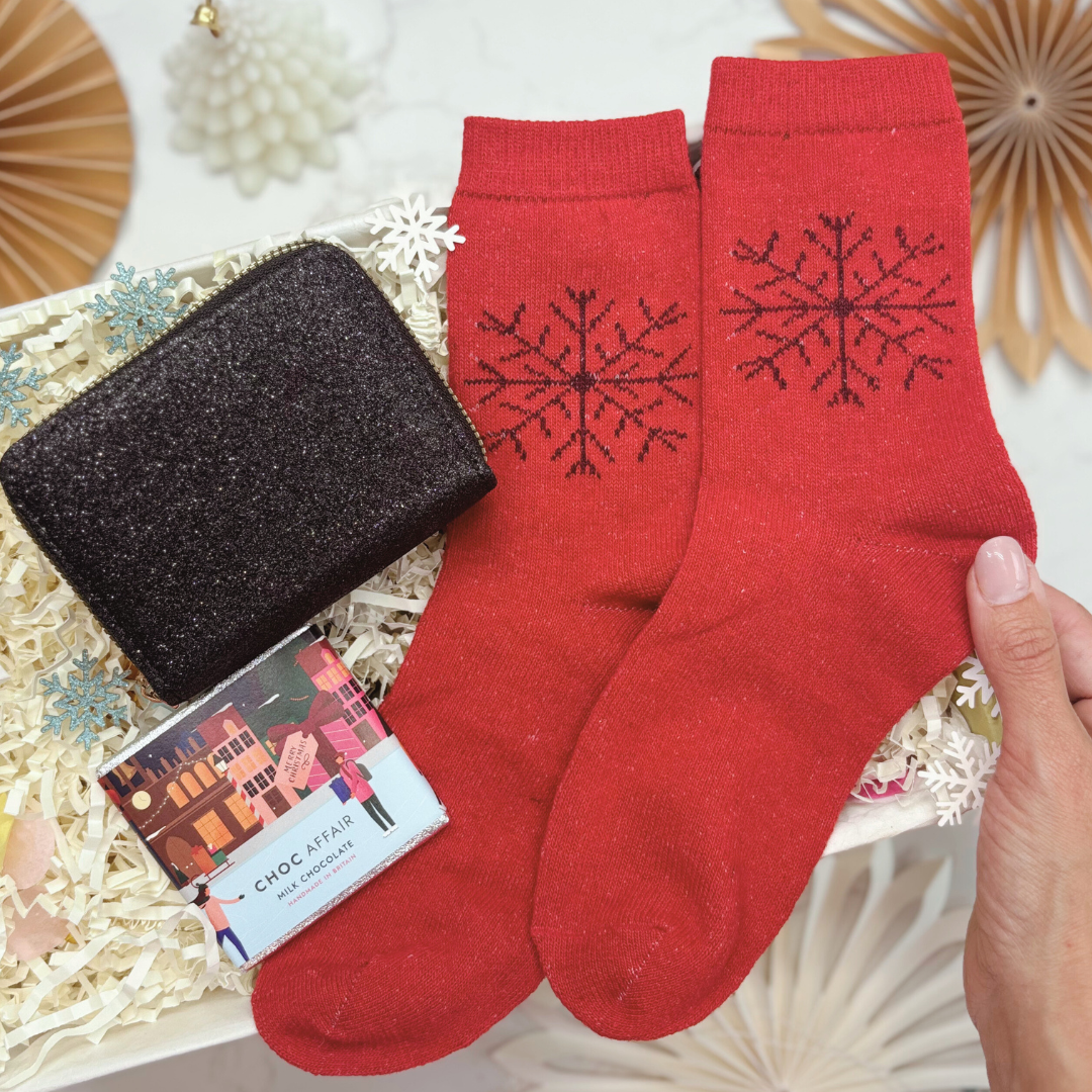 Red socks with snowflake design held by a hand, next to a black glittery purse and chocolate on a decorative giftbox.