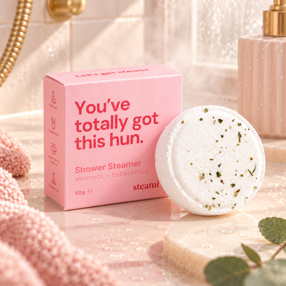Pink shower steamer packaging box next to a round white shower steamer disc with visible green herb flecks, styled on a bathroom counter with a pink towel, gold faucet, and soft warm lighting.