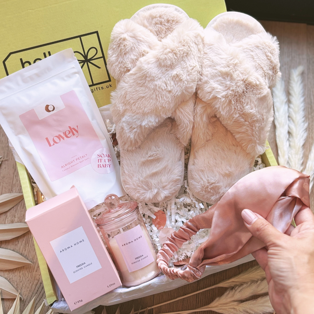 A luxury gift box set containing fluffy slippers, a scented candle in a glass holder, bath salts, and a pink satin hair band.