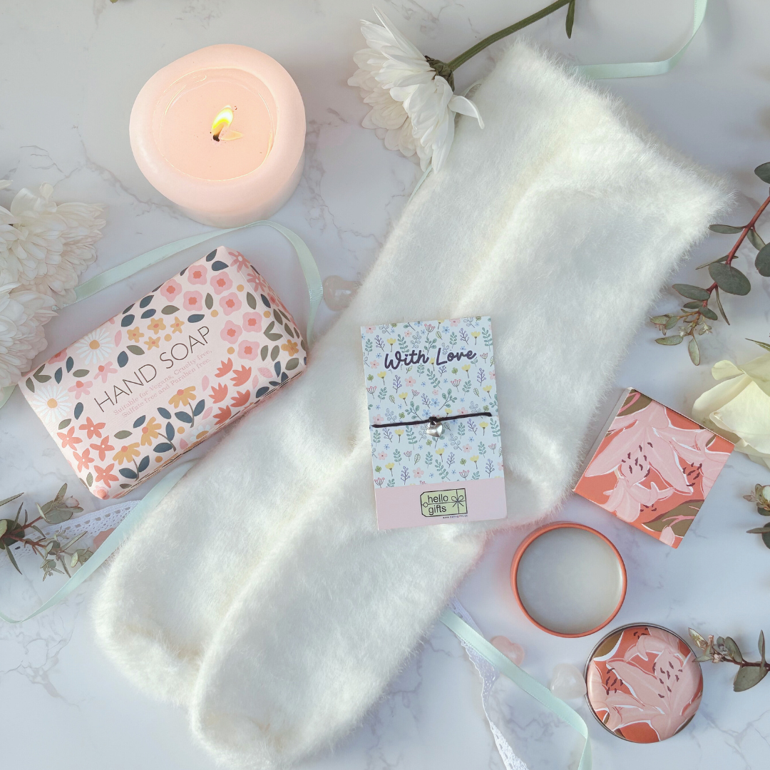 Set of beauty products including a bar of soap, candle, bracelet and fluffy socks on a soft surface with flowers.