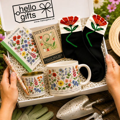 A curated gift box filled with floral-themed items, including a white mug, a small tin, a pencil, a booklet, and a pair of black socks with red flower cuffs, held by hands over a rustic wooden surface with gardening tools and gloves.
