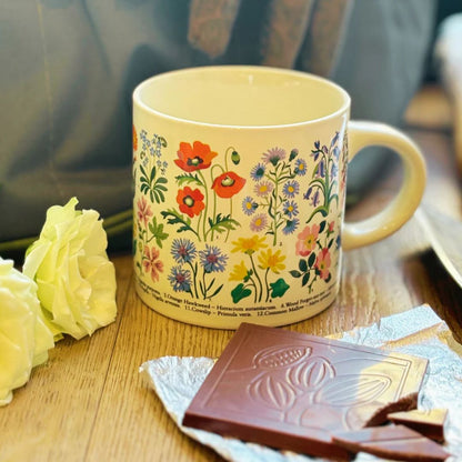 A cream-colored ceramic mug decorated with a colorful wildflower pattern, set on a wooden table next to a yellow rose and a chocolate bar.
