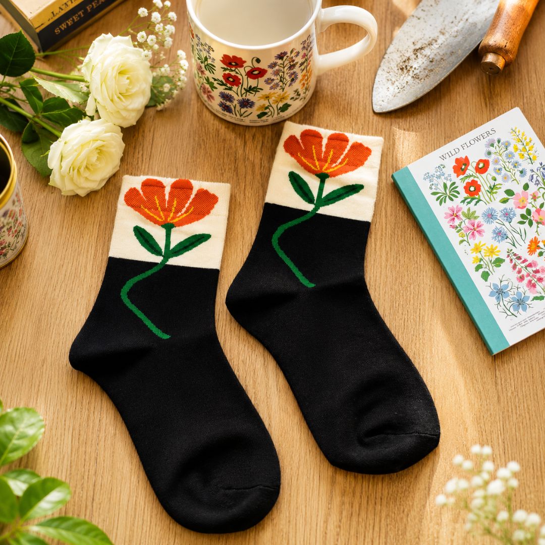A pair of black ankle socks with a white cuff featuring an embroidered orange flower with a green stem, displayed on a wooden tabletop with floral props including a mug, book, and flowers.