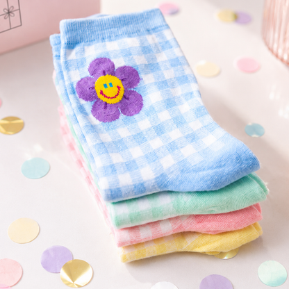 A stack of four checkered ankle socks in pastel colors, featuring a purple flower with a yellow smiley face on the top sock. The socks are styled on a light pink surface with scattered pastel confetti.
