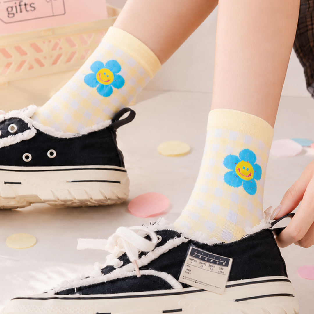 A person wearing pale yellow socks with a white check pattern and a blue smiling flower on the cuff, paired with black canvas low-top sneakers. The background is a soft, light-colored surface with decorative pastel paper pieces.