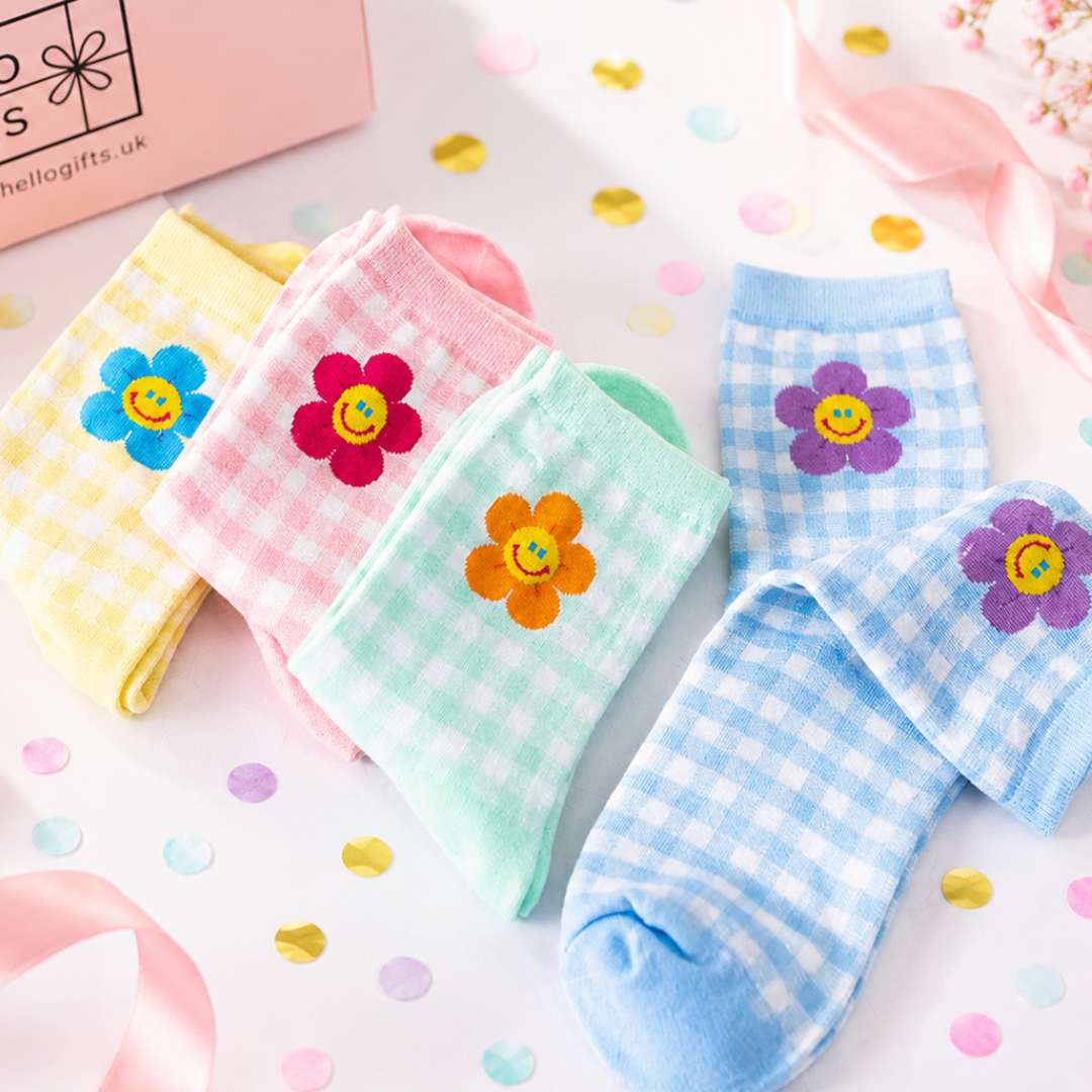 Four pairs of ankle socks arranged on a white surface with confetti and pink ribbon. The socks feature gingham checks in yellow, pink, mint green, and light blue, each decorated with a colorful embroidered flower on the front.
