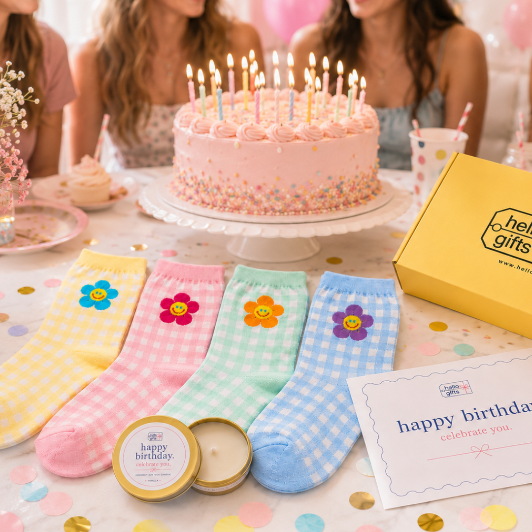A styled birthday celebration scene with a pink frosted cake on a stand, three colorful gingham socks with flower graphics, a yellow Hello Gifts box, a candle tin, and a card on a confetti-covered table, with blurred people seated in the background.
