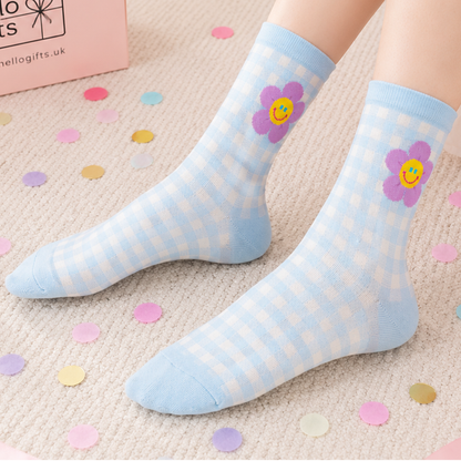 A pair of light blue and white gingham crew socks worn on feet, featuring a purple smiley-face flower design on the upper calf. The socks are shown on a textured beige carpet scattered with pastel confetti, with a pink box partially visible in the top-left corner.