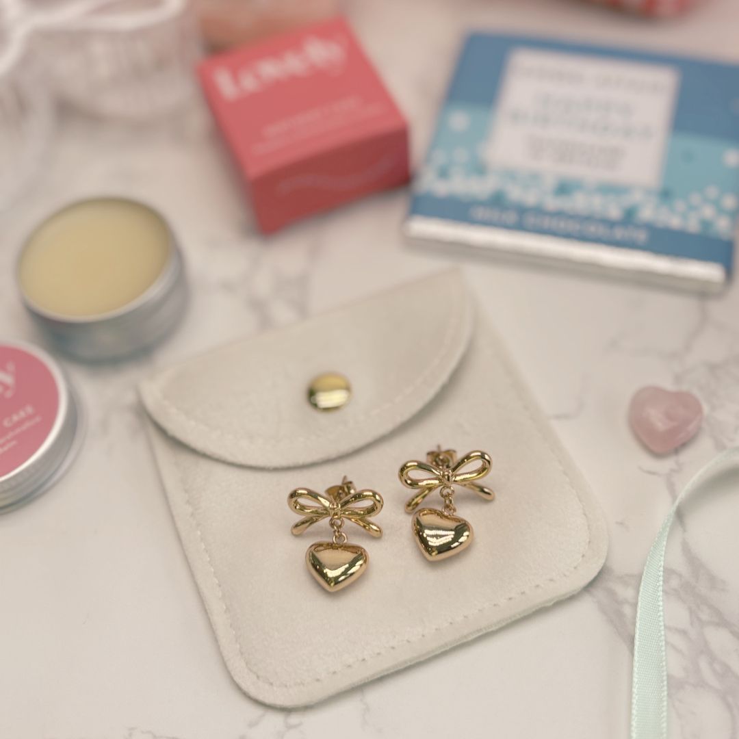 Gold bow and heart-shaped earrings on a white pouch with cosmetic items in the background.