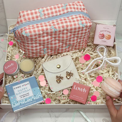 Gift box with a pink gingham make up bag, earrings, and various items on a light background