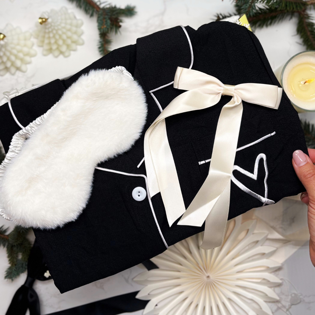 Cotton feel black pyjamas with white  fluffy eye mask, and satin white hair ribbon, held against a decorative giftbox.