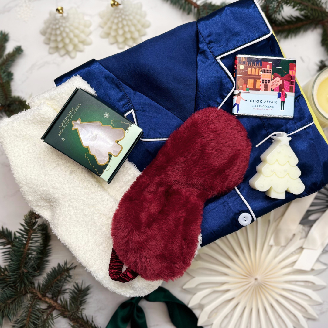 Satin feel festive blue pyjamas with white fluffy socks, christmas tree candle and fluffy eye mask, held against a decorative giftbox.