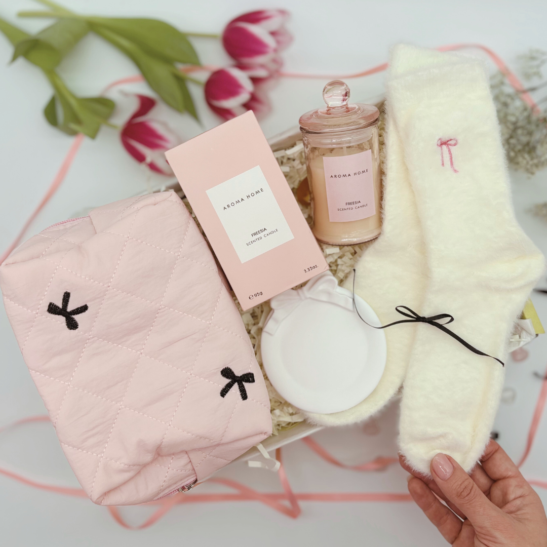 A gift box set containing a blush pink ceramic trinket tray, a pink quilted makeup bag with black bow detail, a scented candle in a glass holder, and fluffy socks with a vintage white and pink bow detail.