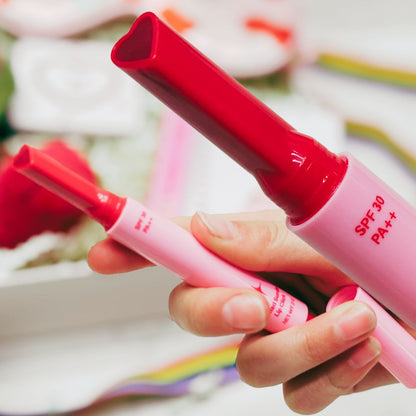 Pink and red lip balm with SPF 30 PA++ held by a hand against a blurred floral background