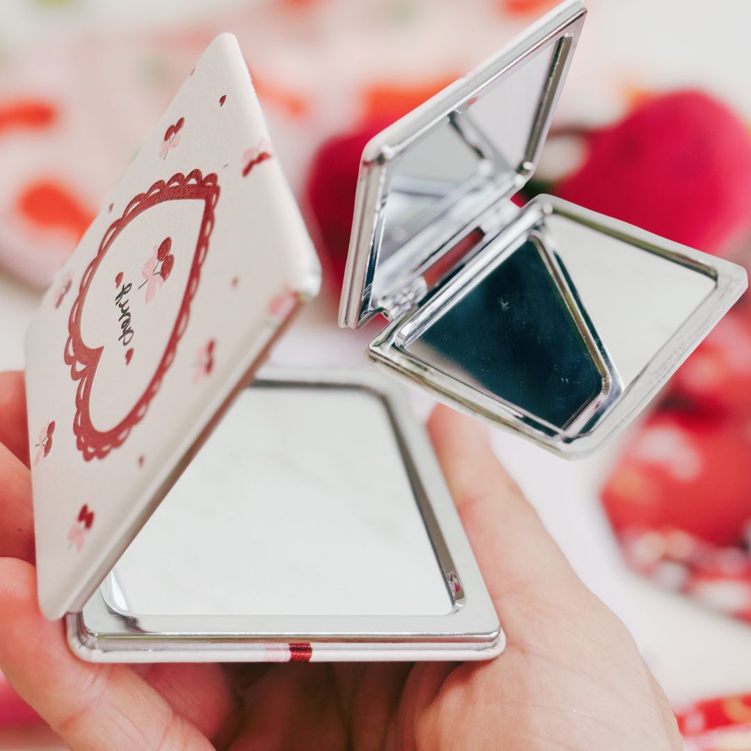 Compact mirror with floral design held by a hand against a blurred floral background