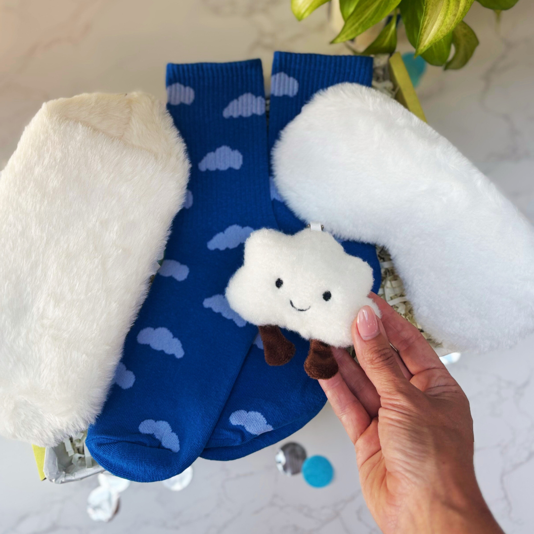 Cloud themed gift box. Cloud toy, socks, fluffy eye mask and make up bag. Blue socks with cloud pattern and a plush cloud toy held by a hand.