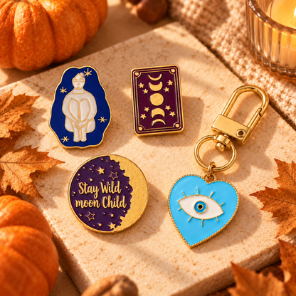 Four celestial-themed enamel pins arranged on a stone surface with autumn props including pumpkins, leaves, and a candle. The pins include a white figure on a blue background, a moon phase design on a purple card, a gold-toned circle reading &