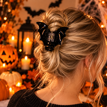 A blonde woman shown from the back wearing a glossy black bat-shaped hair claw clip, set against a warm Halloween background with candles, pumpkins, and string lights.