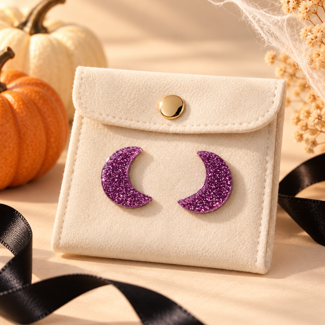 A cream-colored faux leather card holder or small pouch with a gold snap button, featuring two purple glitter crescent moon pins on the front. The pouch is styled with autumn props including pumpkins, dried flowers, and a black ribbon on a beige surface.