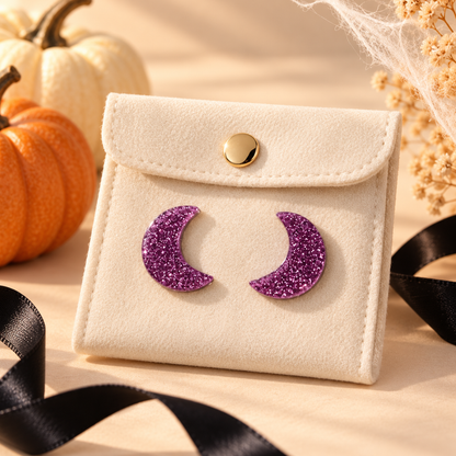 A cream-colored faux leather card holder or small pouch with a gold snap button, featuring two purple glitter crescent moon pins on the front. The pouch is styled with autumn props including pumpkins, dried flowers, and a black ribbon on a beige surface.
