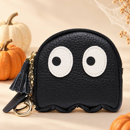 A black ghost-shaped coin purse with large white cartoon eyes, shown with a black tassel keychain and gold ring. The background is a warm beige color with decorative pumpkins and dried flowers.