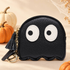 A black ghost-shaped coin purse with large white cartoon eyes, shown with a black tassel keychain and gold ring. The background is a warm beige color with decorative pumpkins and dried flowers.