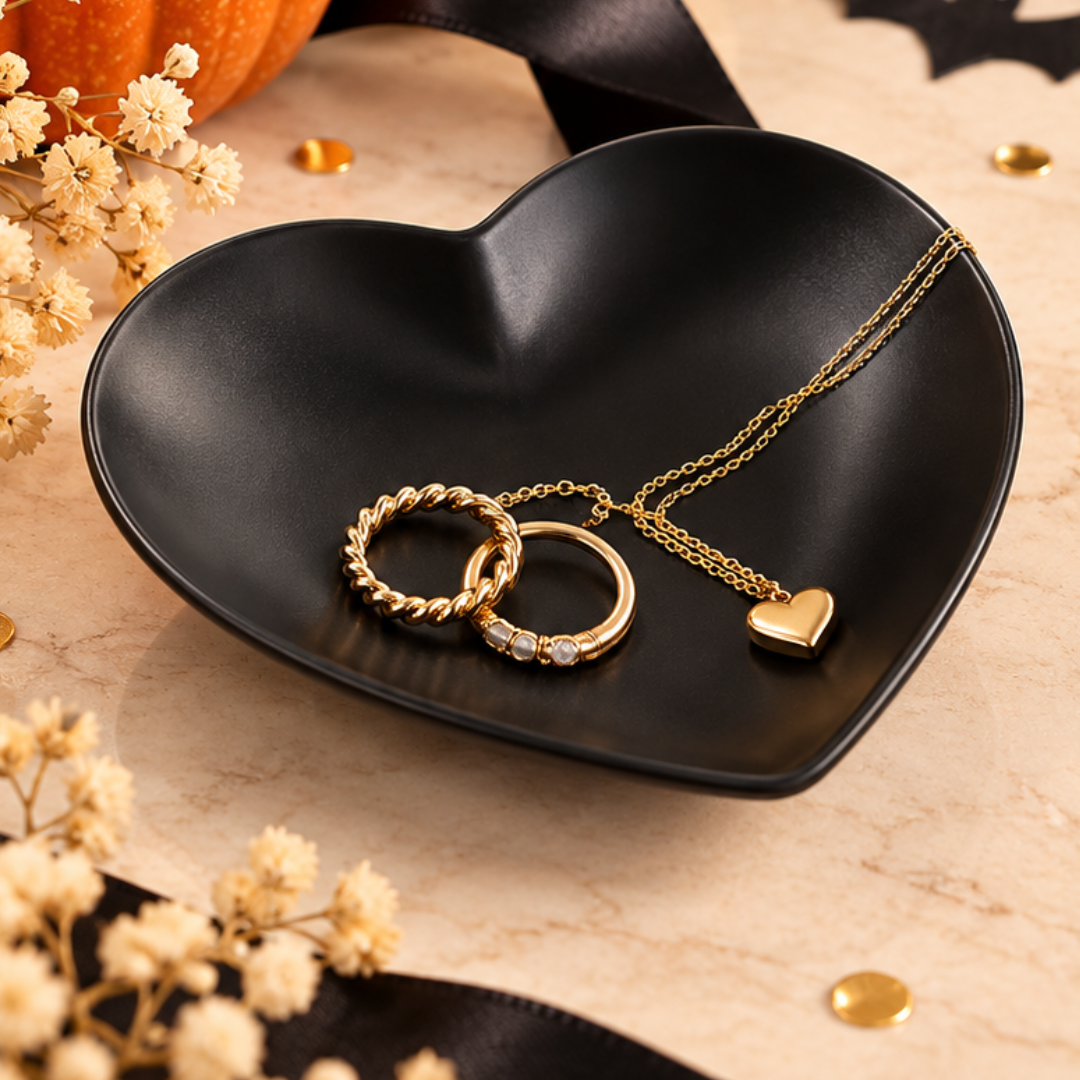 Gold-tone jewelry set featuring two interlocking rings and a heart-shaped pendant on a delicate chain, displayed on a black heart-shaped dish with dried flowers and a pumpkin in the background.