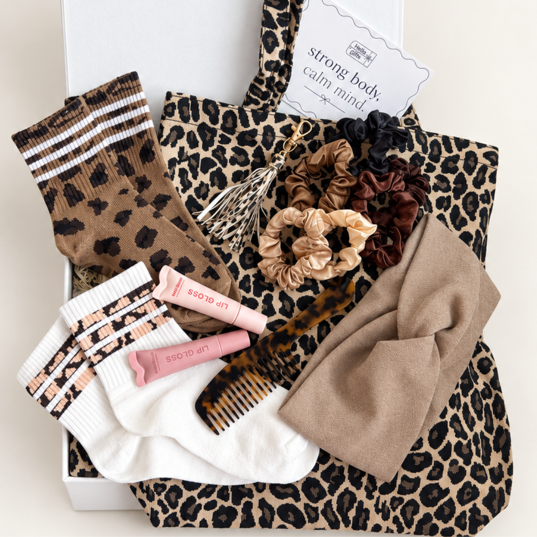 A curated gift box set featuring a leopard print tote bag, a leopard print sock, white socks with brown and pink stripes, a tortoiseshell comb, satin scrunchies in beige, black, and brown, and two pink lip balm tubes, all arranged in a white box with a card that reads &