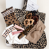 A curated gift box set featuring a leopard print tote bag, a leopard print sock, white socks with brown and pink stripes, a tortoiseshell comb, satin scrunchies in beige, black, and brown, and two pink lip balm tubes, all arranged in a white box with a card that reads &