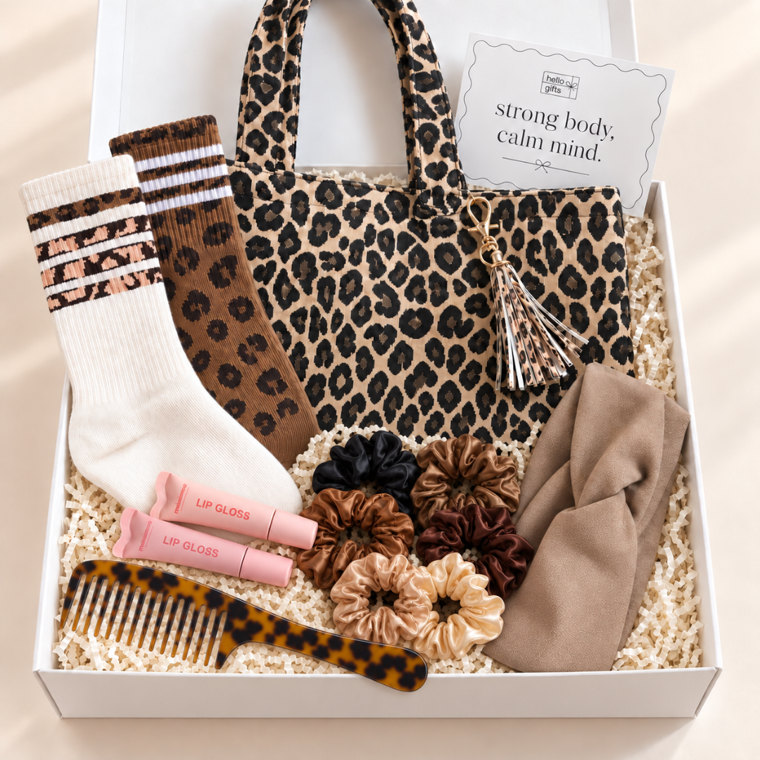 A curated gift box set in a white box filled with beige paper shred, featuring a leopard-print tote bag, a cream and brown striped sock, a brown patterned sock, pink lip gloss tubes, multiple fabric scrunchies, a tortoiseshell hairbrush, and a beige headband. A card inside the box reads &