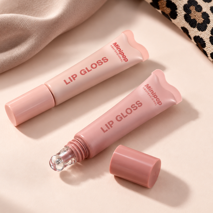 Two pink lip gloss tubes from Minijop on a light beige surface, one upright and one lying down with its cap removed to show the clear roller ball, styled with a beige fabric and a leopard print corner.