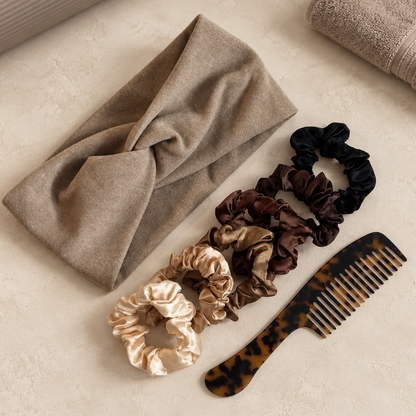 A set of six satin scrunchies in beige, brown, and black, displayed alongside a tortoiseshell hair comb and a folded beige scarf on a textured beige surface.