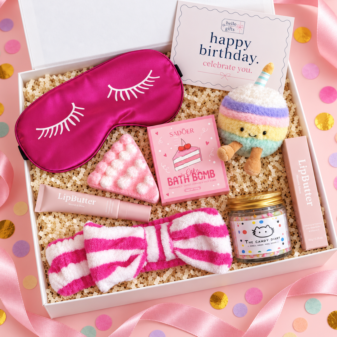 A curated beauty gift box set in a white box filled with shredded paper, featuring a hot pink satin sleep mask with white eyelash embroidery, a pink striped plush bow headband, a pink bath bomb, a pink lip butter tube, a small jar candle, and a pastel plush toy, all arranged on a pink confetti background with a card reading &
