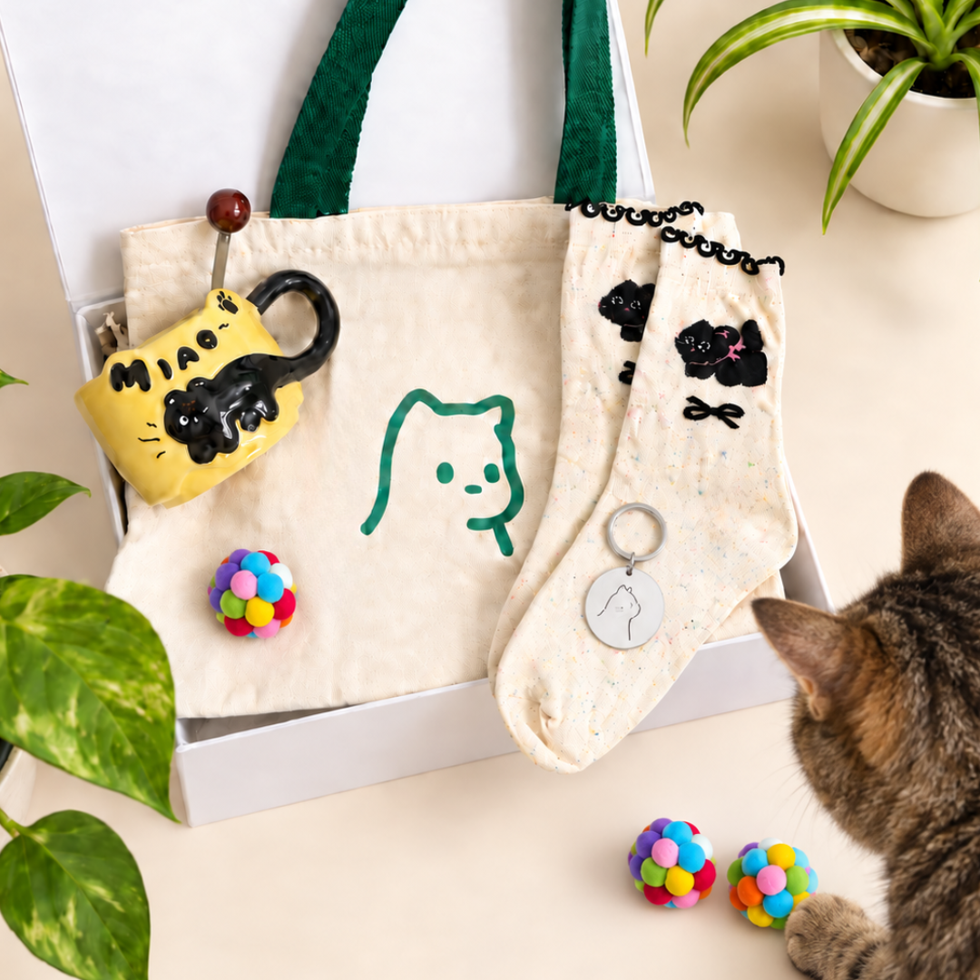 A styled product display of a cat-themed gift set inside an open white box, featuring a cream canvas tote bag with a green cat face, a yellow cat-shaped keychain, a pair of cream socks with black cat illustrations, two colorful pom-pom balls, and a tabby cat partially visible at the bottom right.