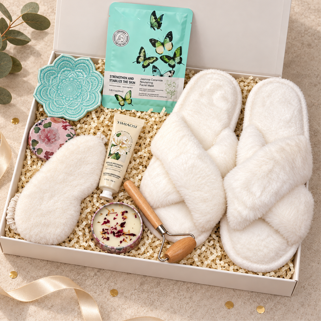 A curated gift box set featuring fluffy white slippers, a white eye mask, a tube of hand cream, a butterfly-themed facial mask packet, a teal flower-shaped candle, and a rose petal roller, all arranged in a white box filled with shredded paper.