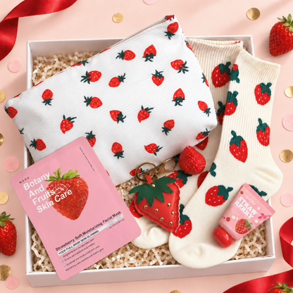 A strawberry-themed gift set in a white box, featuring a white pouch and a pair of white socks with red strawberry prints, a pink facial mask packet, a strawberry-shaped keychain, and a pink strawberry hand cream tube, arranged on a pink background with confetti and ribbon.
