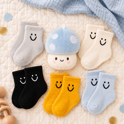 Six pairs of baby socks arranged on a quilted cream blanket with a soft blue cloth and small gold confetti. The socks come in gray, cream, black, yellow, and light blue, each with a simple black smiley face.